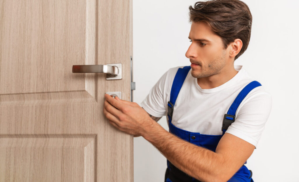 Professional Repair Service. Portrait of focused young male locksmith workman in blue uniform installing door knob. Installation of a lock on the front wooden entrance door at home or office workplace